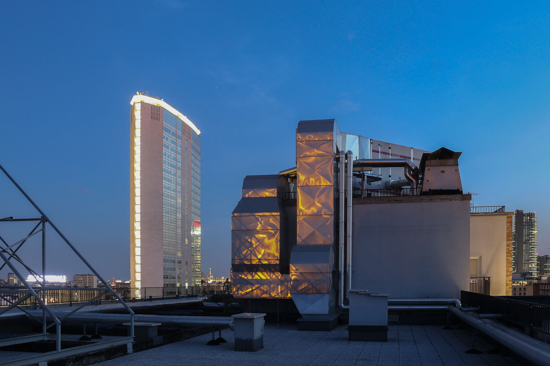 Il Pirellone illuminato al blue hour, visto da un rooftop con impianti tecnici in primo piano.