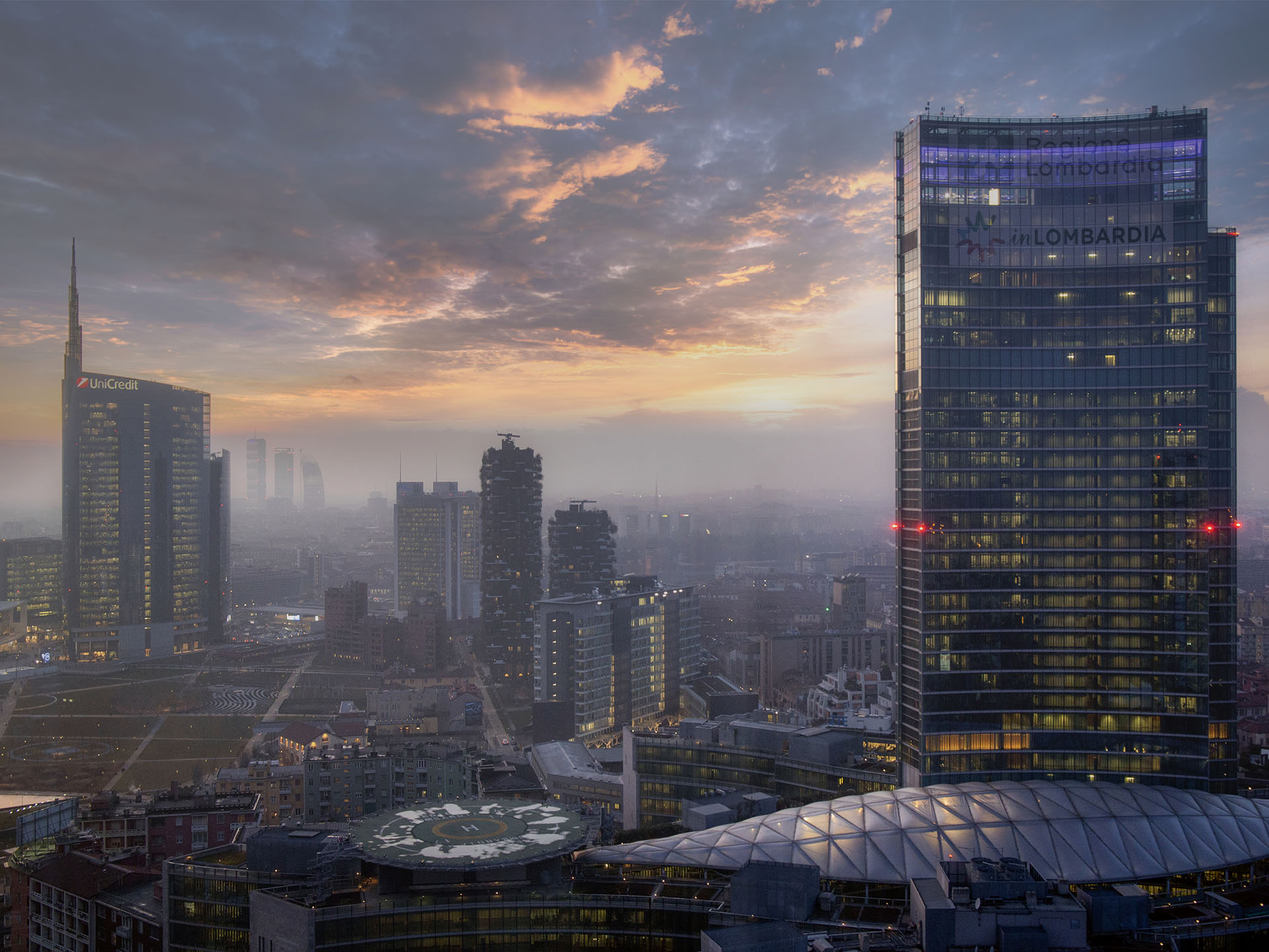 Palazzo Lombardia in primo piano tra la nebbia all'alba, con la Torre Unicredit sullo sfondo.