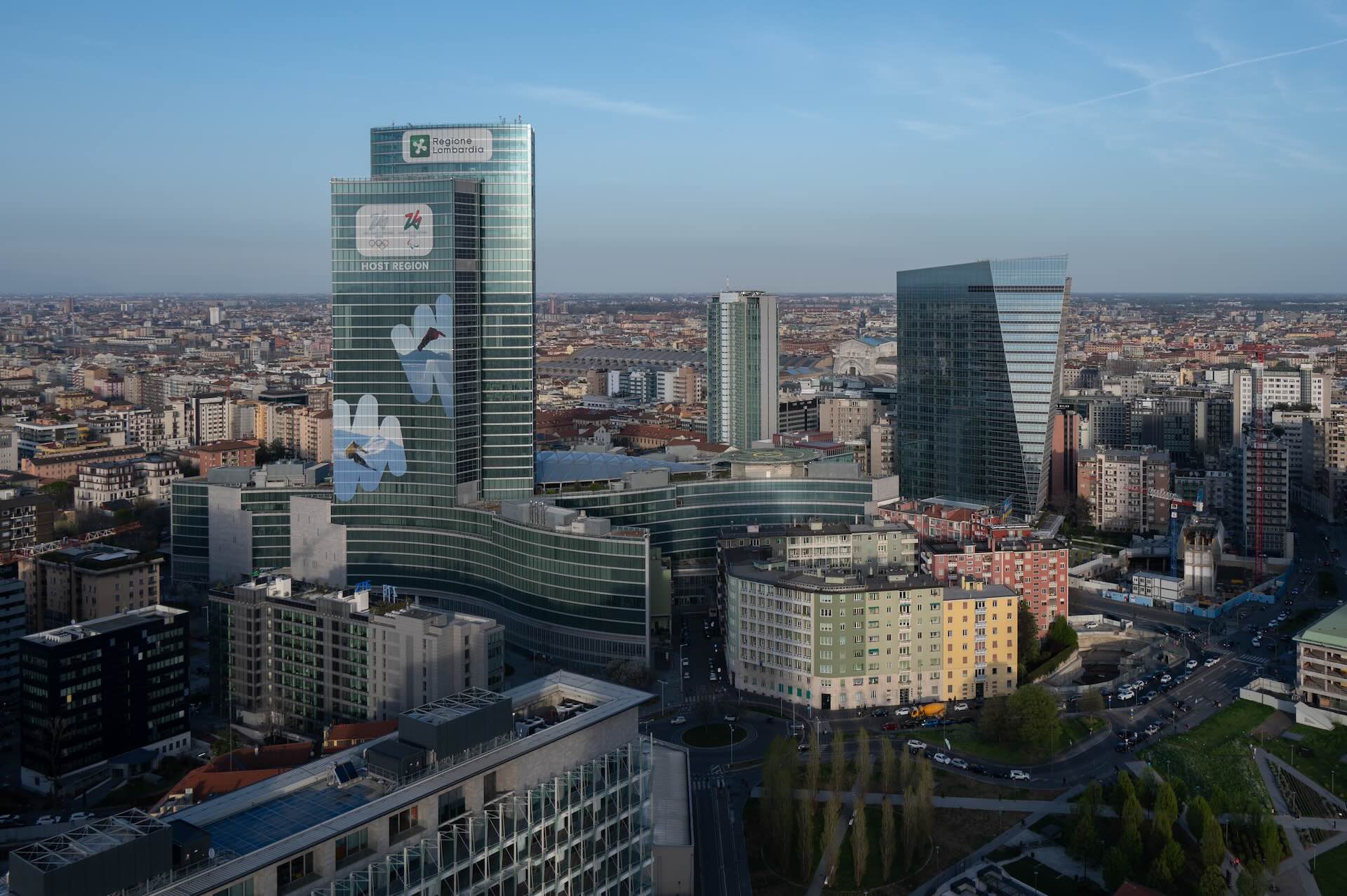 Vista aerea diurna di Palazzo Lombardia con il banner 'Host Region' delle Olimpiadi Milano-Cortina 2026.