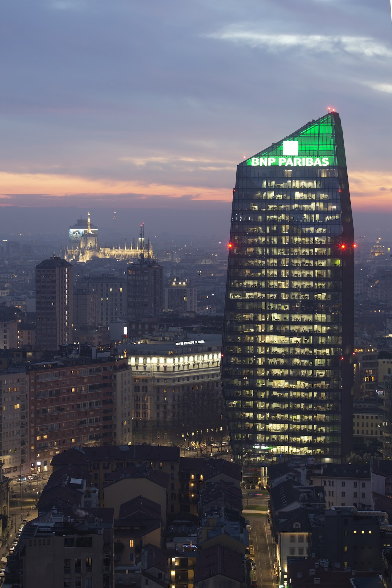 La Torre Diamante al crepuscolo con l'insegna BNP Paribas illuminata di verde, il Duomo silhouette e l'Hotel Principe di Savoia in primo piano.