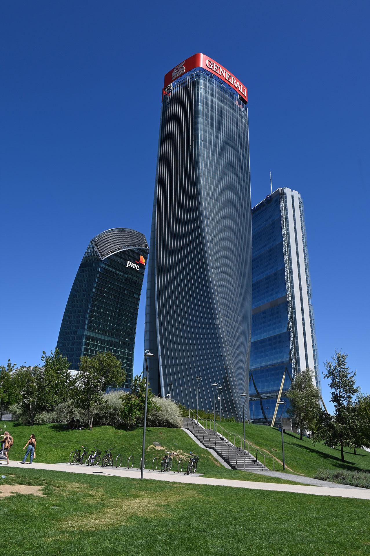 Le Tre Torri di CityLife — PwC, Generali e Allianz — viste dal parco, con persone e biciclette in primo piano.