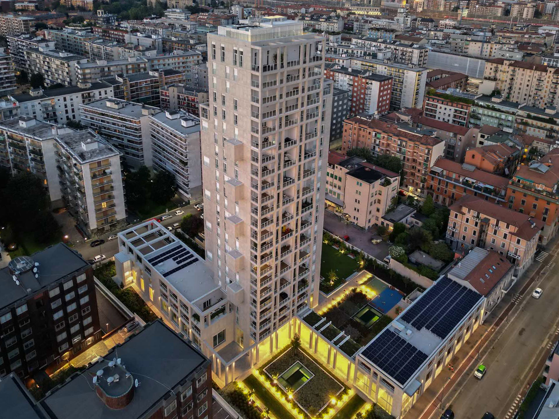 Vista aerea al crepuscolo di Torre Milano, con il giardino pensile illuminato e i pannelli solari sul tetto adiacente.