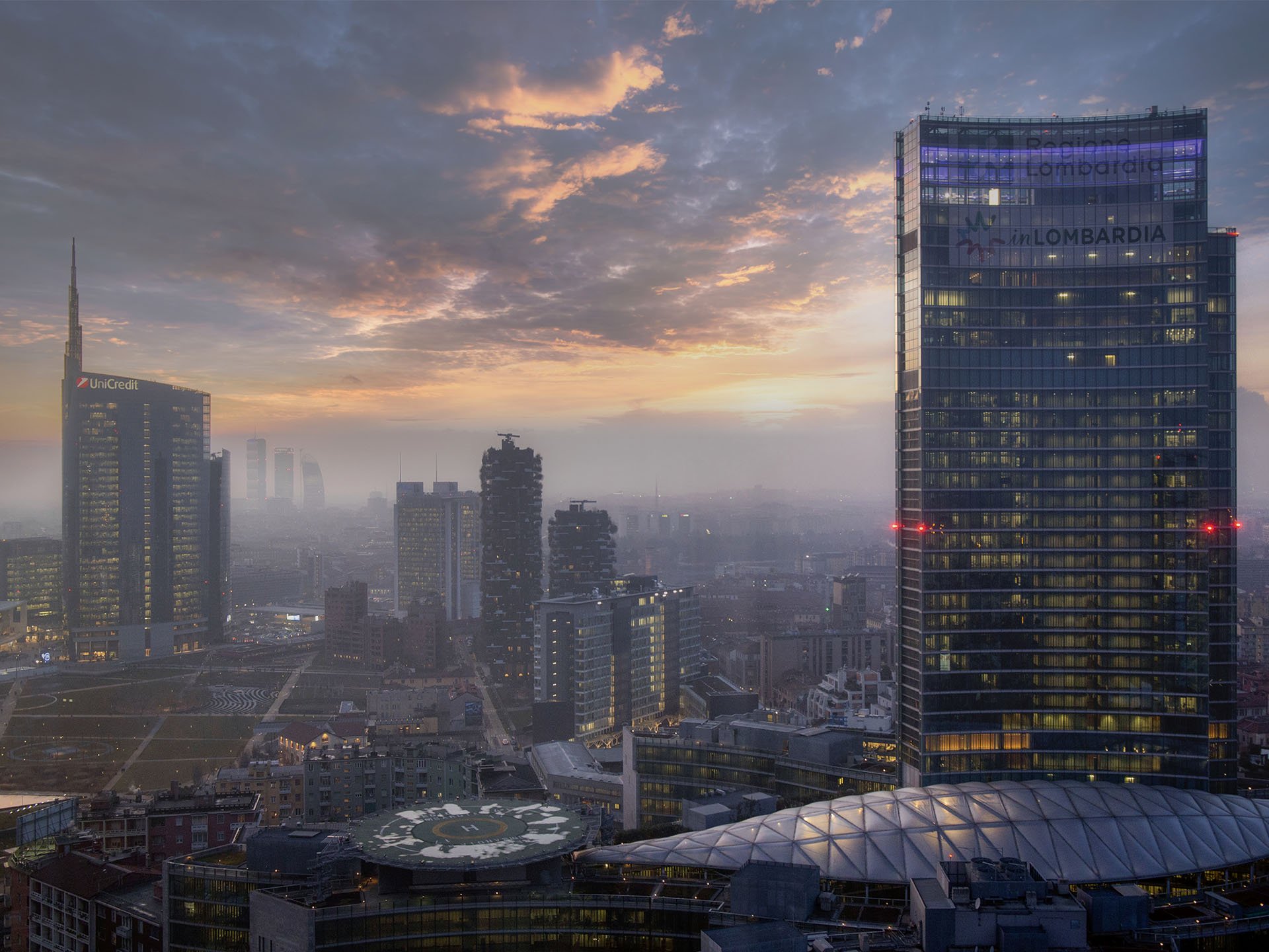 Palazzo Lombardia in primo piano tra la nebbia all'alba, con la Torre Unicredit sullo sfondo.