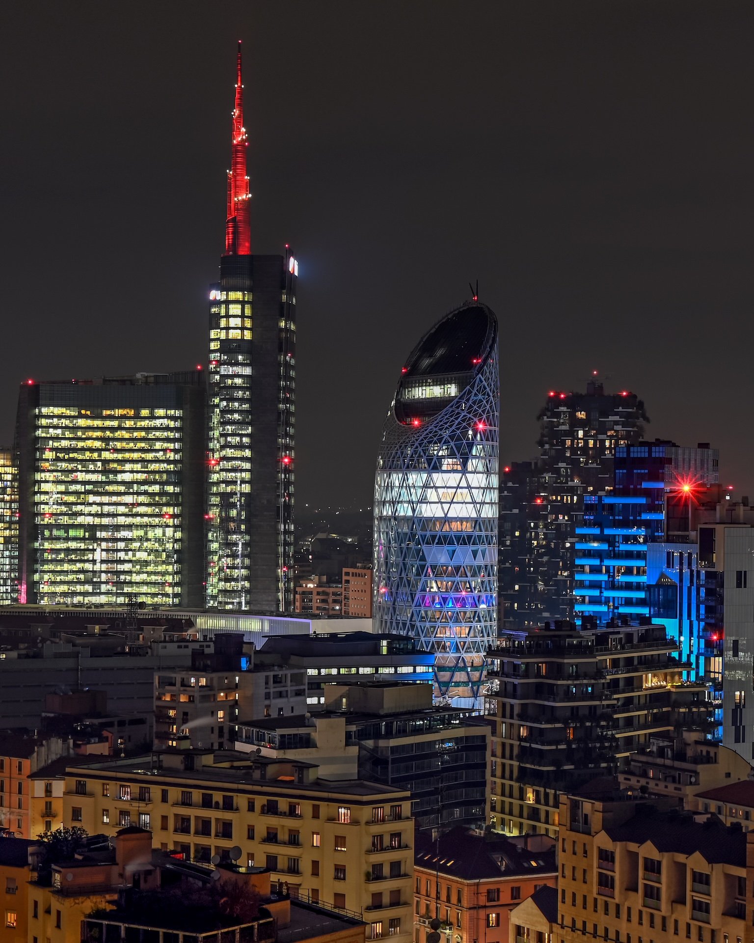 La guglia della Torre Unicredit illuminata di rosso di notte, con il Bosco Verticale e la Torre Diamante sullo sfondo.